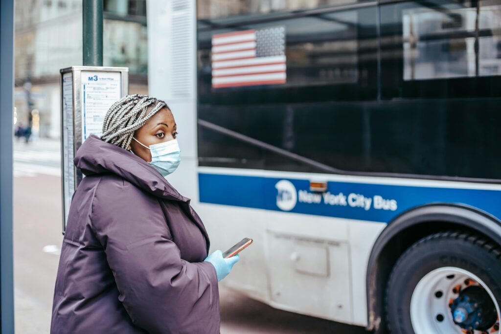 Woman with mask and mobile device waiting at New York City bus stop during pandemic.